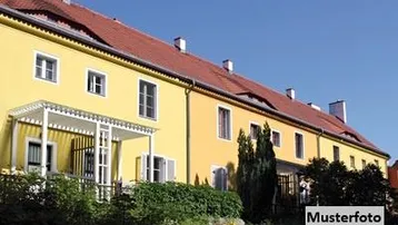 Gelbes Gebäude mit rotem Ziegeldach, einem kleinen Balkon und grüner Bepflanzung unter blauem Himmel.