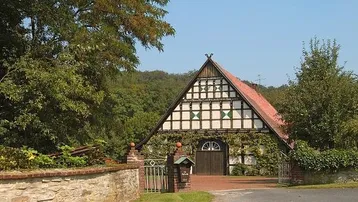 Traditionelles Fachwerkhaus mit markantem Giebeldach, umgeben von üppiger Vegetation und einem Steinzaun.