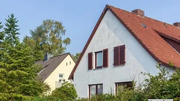 Weißes Haus mit rotem Satteldach und braunen Fensterläden, umgeben von grünen Büschen unter blauem Himmel.