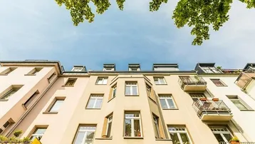 Fassade eines Mehrfamilienhauses mit Balkonen und Blick auf den Himmel durch Baumkronen.
