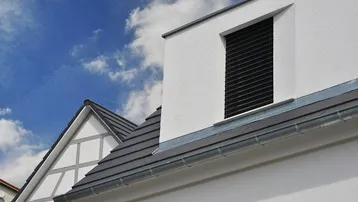 Nahaufnahme einer weißen Hausfassade mit Dachgaube und dunklem Ziegeldach unter blauem Himmel.