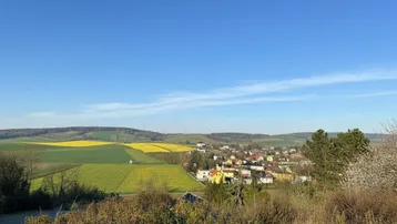Weitläufiger Blick über eine ländliche Landschaft mit gelben Feldern und einem charmanten Dorf in der Ferne unter blauem Himmel.