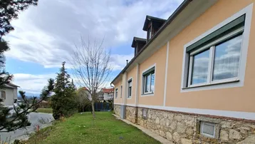 Hausfassade mit beiger Putzfassade, Steinsockel und grünen Fensterläden, umgeben von einem gepflegten Rasen.