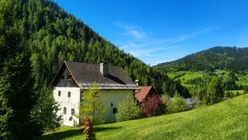 Traditionelles Haus in einer grünen Berglandschaft unter blauem Himmel mit viel Wald.