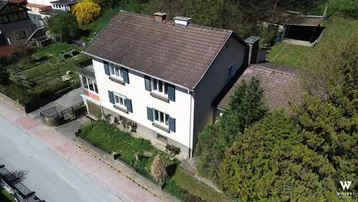 Luftaufnahme eines traditionellen Einfamilienhauses mit Satteldach, blauen Fensterläden und umliegendem Garten.