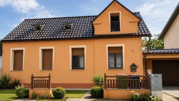 Einfamilienhaus mit oranger Fassade, dunklem Ziegeldach und gepflegtem Vorgarten unter blauem Himmel.
