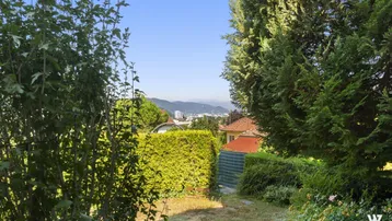 Gartenbereich mit Hecken, Bäumen und Fernblick auf die Stadt und Berge.