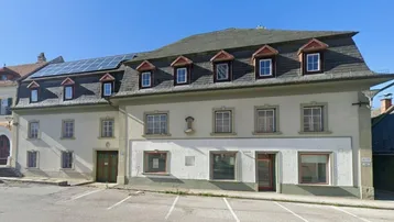 Altes Gebäude mit Mansarddach und Solarpaneelen auf dem Dach, umgeben von Parkplätzen.