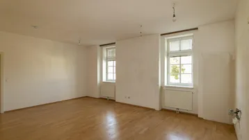 Geräumiger, unmöblierter Wohn-Essbereich mit hellem Parkettboden und zwei großen Fenstern.