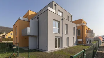 Modernes Mehrfamilienhaus mit grauer und orangefarbener Fassade, umgeben von Grünflächen und einem Zaun.