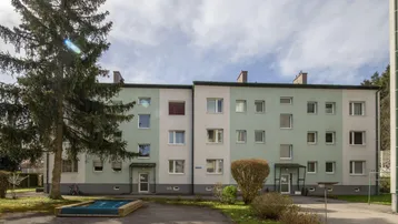 Mehrfamilienhaus mit hellgrüner und weißer Fassade, umgeben von Grünflächen und Bäumen.