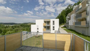 Geräumiger Balkon mit Fliesenboden und weitem Blick auf die grüne Landschaft und umliegende Gebäude.