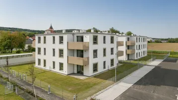 Moderne Neubauten mit weißer Fassade und Balkonen, umgeben von Grünflächen und Parkplätzen unter blauem Himmel.