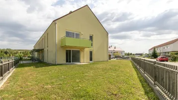 Gelbes Mehrfamilienhaus mit grünem Balkon und Rasenfläche unter bewölktem Himmel.