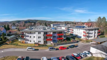 Mehrere moderne Wohngebäude mit roten Balkonen und grüner Umgebung unter blauem Himmel.