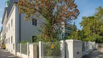 Graues Mehrfamilienhaus mit moderner Architektur und gepflegter Außenanlage unter einem großen Baum.