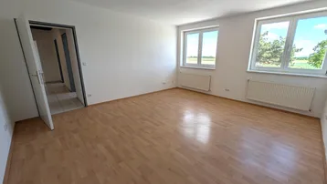 Helles, leeres Zimmer mit Laminatboden und Blick ins Grüne durch große Fenster.