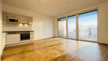 Heller offener Wohnbereich mit integrierter Küchenzeile, Parkettboden und Zugang zu einem Balkon.