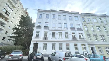 Weißes Mehrfamilienhaus mit mehreren Stockwerken und parkenden Autos auf der Straße.