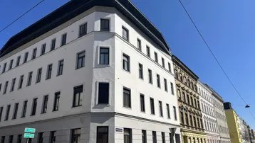 Eckgebäude mit weißer Fassade und dunklen Fenstern unter klarem blauem Himmel.