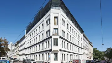 Modernes Eckgebäude mit weißer Fassade und Dachterrasse unter blauem Himmel.