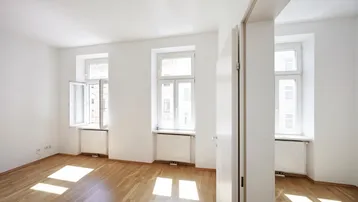 Helles, leeres Zimmer mit Parkettboden und mehreren Fenstern, das viel Tageslicht hereinlässt.