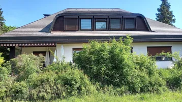 Hausfassade mit markantem Mansarddach, Gaubenfenstern und üppiger Bepflanzung im Vorgarten.