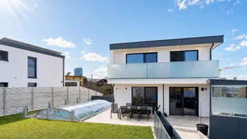 Modernes Einfamilienhaus mit großem Garten, Terrasse und überdachtem Pool unter strahlend blauem Himmel.