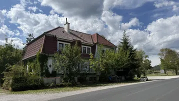 Altes Haus mit Mansarddach und grüner Fassade, umgeben von Bäumen und einer Straße.