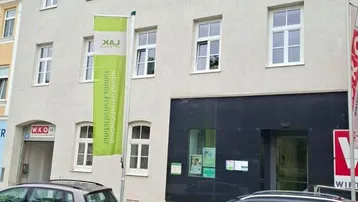 Ansicht der Gebäudefassade mit mehreren Fenstern, einem Eingangsbereich und parkenden Autos auf der Straße.