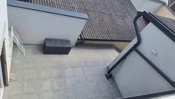 Blick von oben auf eine gepflasterte Terrasse mit einem Abstellkasten und angrenzenden Dächern.