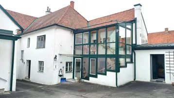 Weißes Gebäude mit roten Ziegeldächern und einem angebauten Wintergarten mit grünen Fensterrahmen.