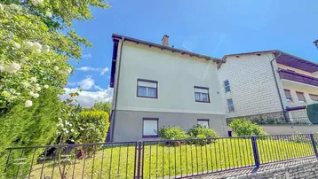 Helles Einfamilienhaus mit gepflegtem Vorgarten und Zaun unter blauem Himmel an einem sonnigen Tag.