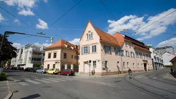 Traditionelles Gebäude an einer Straßenecke mit hellrosa Fassade und roten Ziegeldächern unter blauem Himmel.
