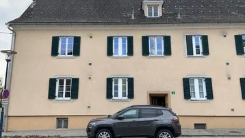 Traditionelles Gebäude mit beiger Fassade, grünen Fensterläden und einem geparkten Auto davor.