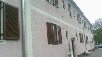 Fassade eines Wohngebäudes mit hellrosa Putz, braunen Fensterläden und einem Regenrohr.