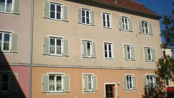 Mehrstöckiges Wohngebäude mit beigefarbener Fassade, roten Dachziegeln und grünen Fensterläden.