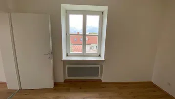 Schlafzimmer mit Laminatboden, Fenster und Blick auf die umliegenden Dächer und Berge.