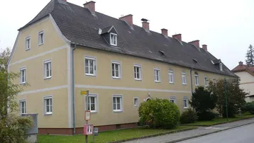 Gut erhaltenes Mehrfamilienhaus mit Mansarddach, weißen Fenstern und grüner Umgebung.