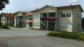 Mehrfamilienhaus mit beiger Fassade, Satteldach und Balkonen mit Blumenkästen sowie Außenparkplätzen und Grünanlage.