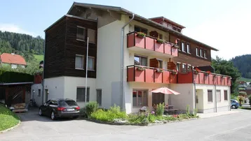 Mehrfamilienhaus mit Balkonen, Parkplätzen und einer kleinen Terrasse im Grünen, umgeben von Hügeln.