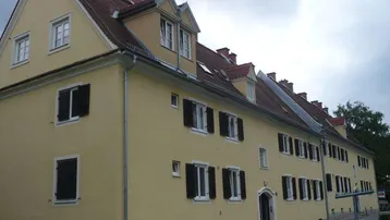 Gelbe Fassade eines Mehrfamilienhauses mit roten Ziegeldächern und dunklen Fensterläden.
