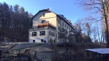 Historisches Gebäude auf einem Hügel, umgeben von Bäumen, mit Balkonen und weitem Blick ins Grüne.