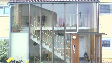 Gebäudefassade mit verglastem Treppenhaus, Balkonen und Eingangsbereich, umgeben von Grünflächen.