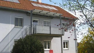 Gebäudeansicht mit Balkon, Solarpaneelen auf dem Dach und grüner Umgebung.