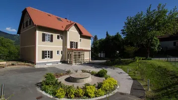 Modernes Gebäude mit Satteldach, umgeben von Grünflächen und einem gepflegten Vorgarten mit Zufahrt.