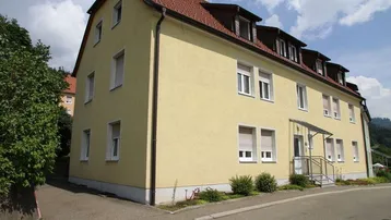 Mehrfamilienhaus mit gelber Fassade, rotem Ziegeldach und mehreren Fenstern mit Rollläden.