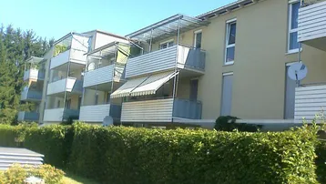 Mehrfamilienhaus mit mehreren Balkonen und grüner Hecke im Vordergrund.