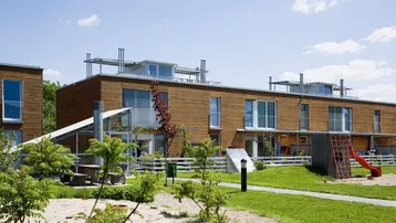 Moderne Reihenhäuser mit Holzfassade und Spielplatz im Gemeinschaftsgarten unter blauem Himmel.