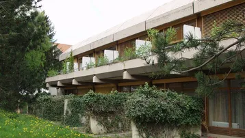 Gebäude mit mehreren Balkonen und Holzjalousien, umgeben von üppigem Grün und einem Rasen.